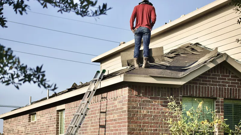 Roofer standing on a residential roof inspecting storm-damaged shingles